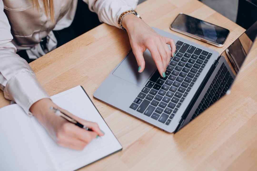 Une femme prend des notes de la main droite pendant que sa main gauche pianote sur un ordinateur portable